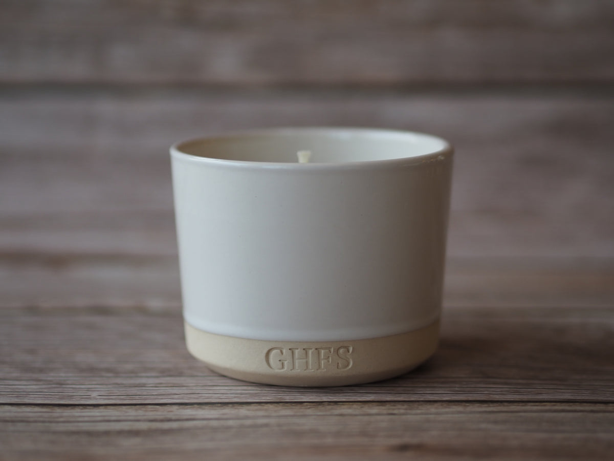 A ceramic candle in a cylindrical white and beige pot with the brand name 'GHFS' embossed on the base, set on a rustic wooden surface.